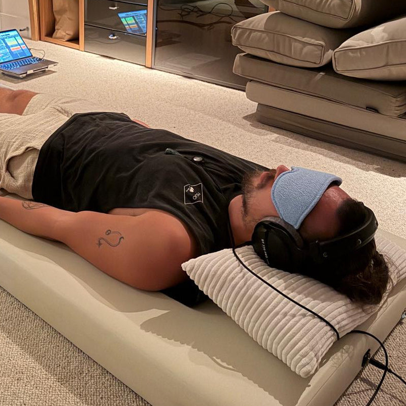 Jake Delmonte lying on an Onizion sound bed in a living room, wearing studio headphones and an eye mask